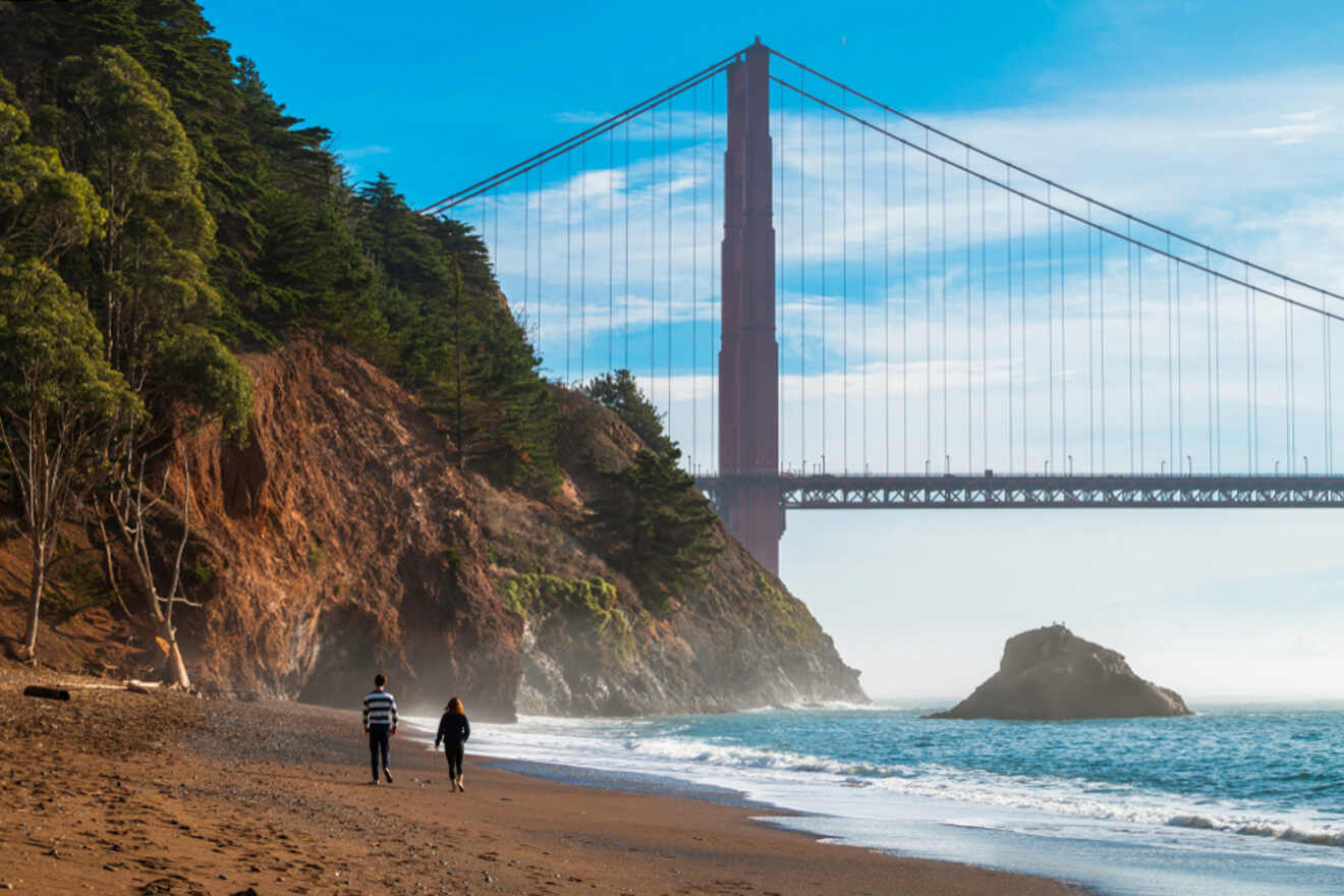 two people walking on a beach near the ocean under a bridge