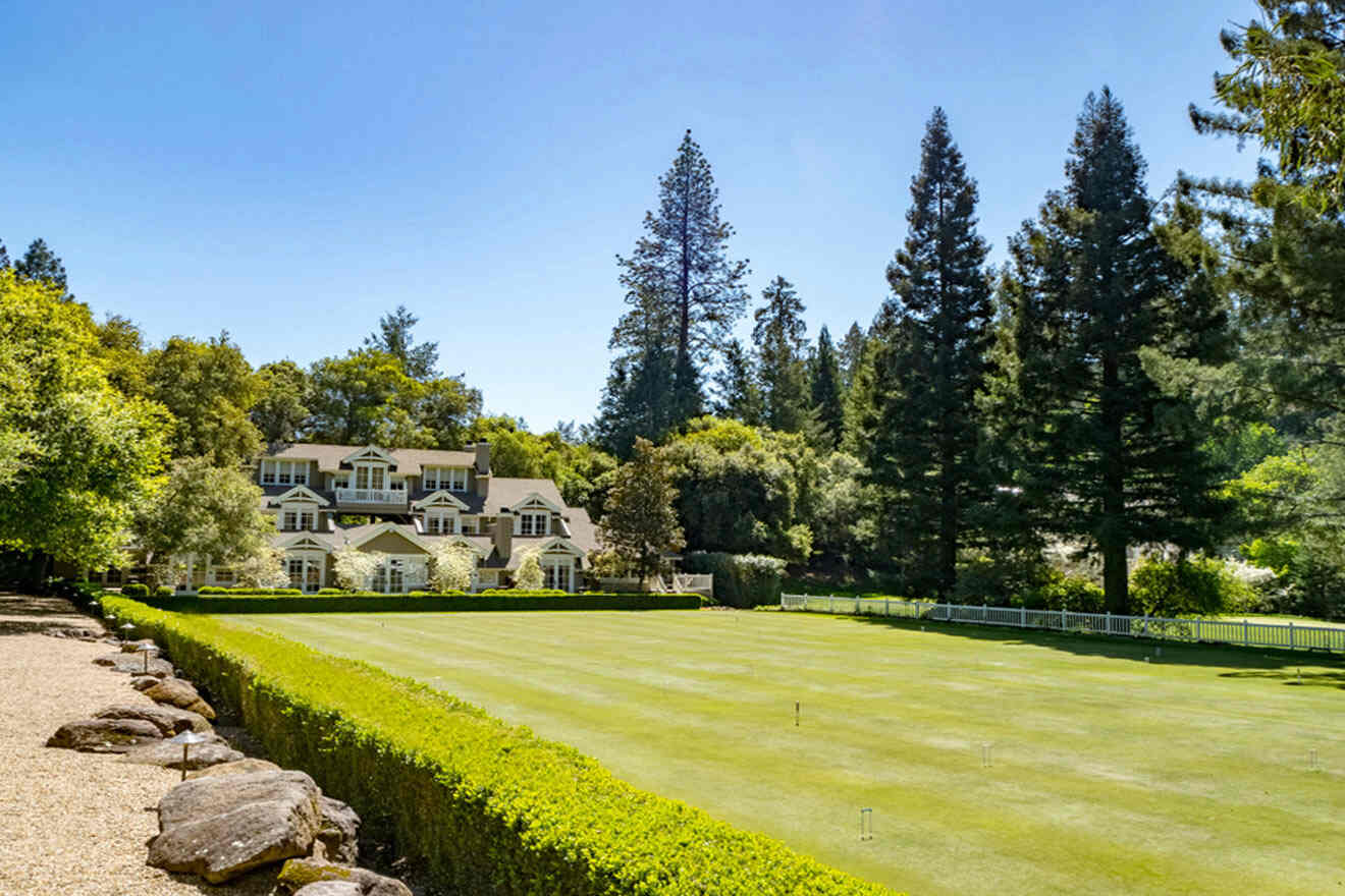 Large house with a light exterior behind a manicured lawn and hedges, surrounded by tall trees under a clear blue sky.