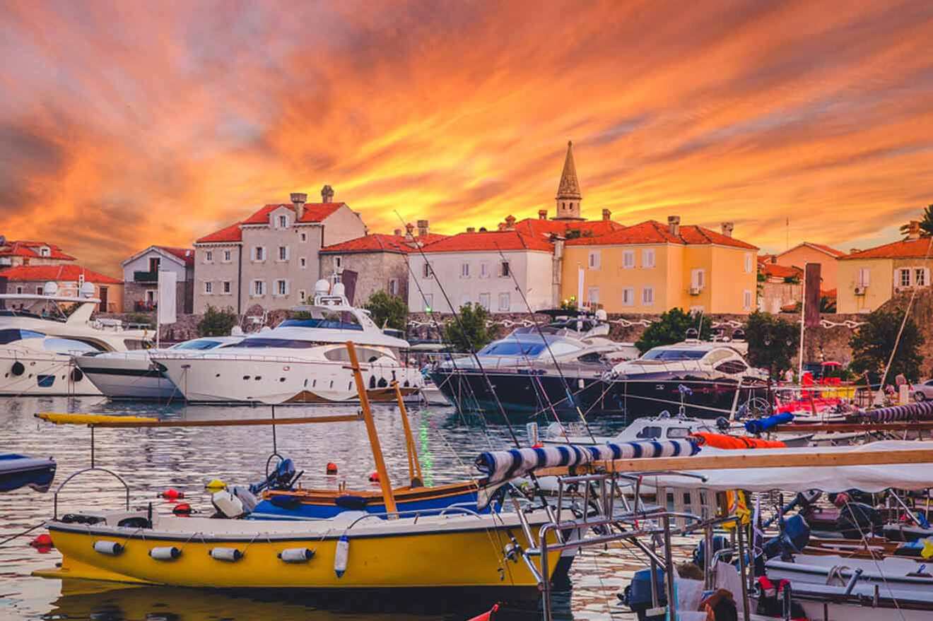 Sunset over a harbor with several yachts and small boats docked. The sky is illuminated with vibrant orange and yellow hues, with buildings and a church tower visible in the background.