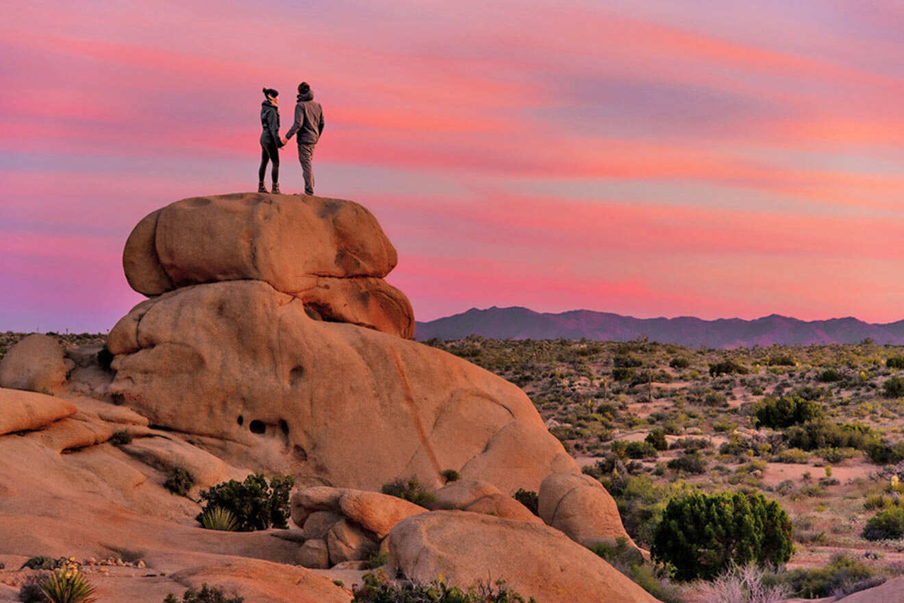 two people standing on top of a large rock looking at a stunning pink sky sunset