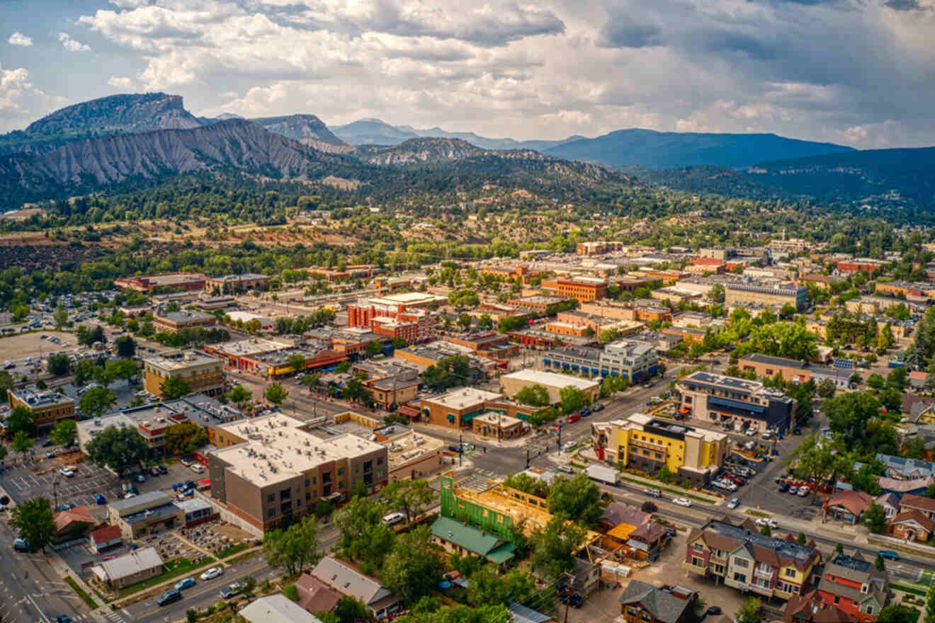 Aerial view of a town with numerous buildings and streets, surrounded by hills and mountains under a partly cloudy sky.