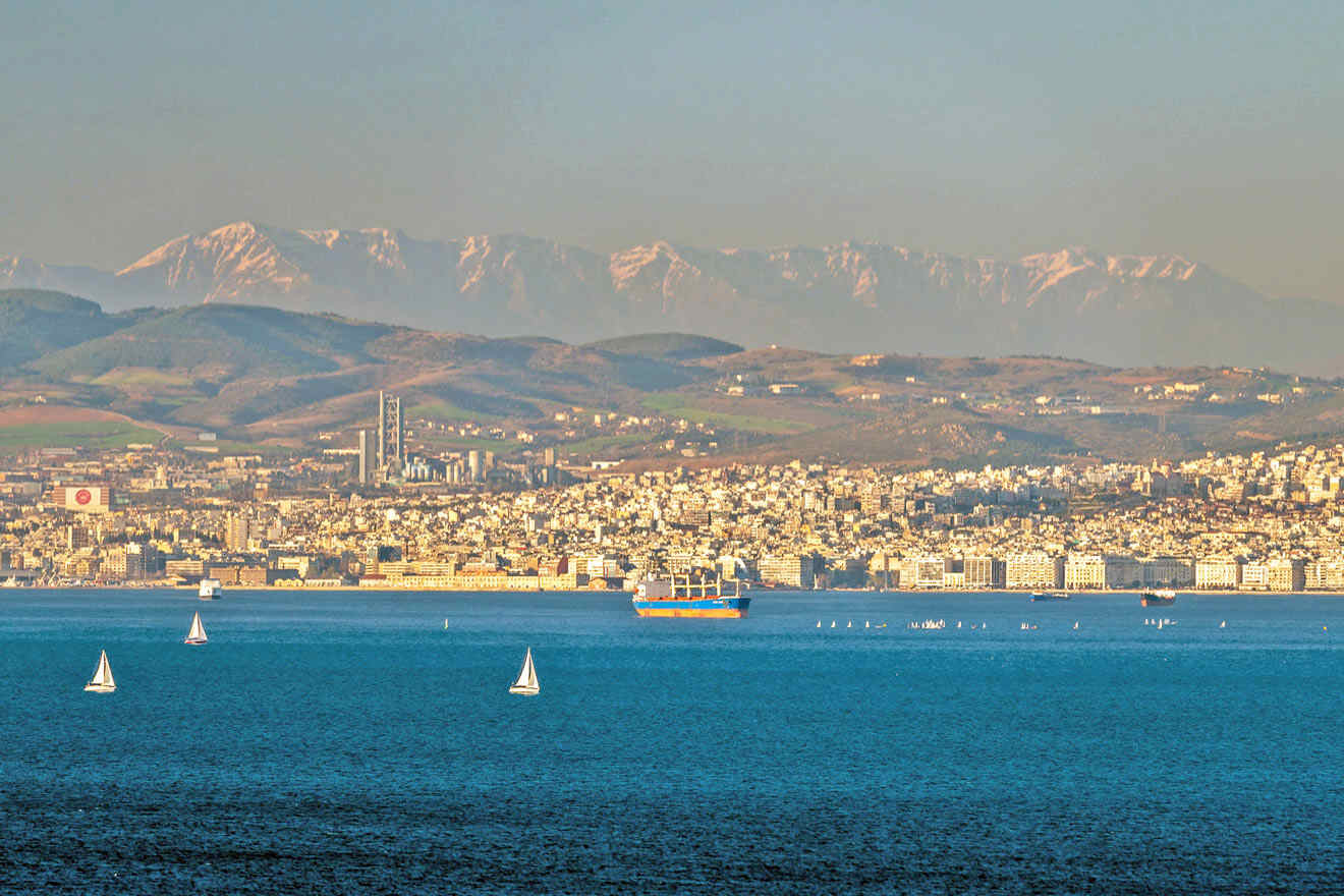 Coastal cityscape with buildings, mountains in the background, and several sailboats on a calm blue sea.