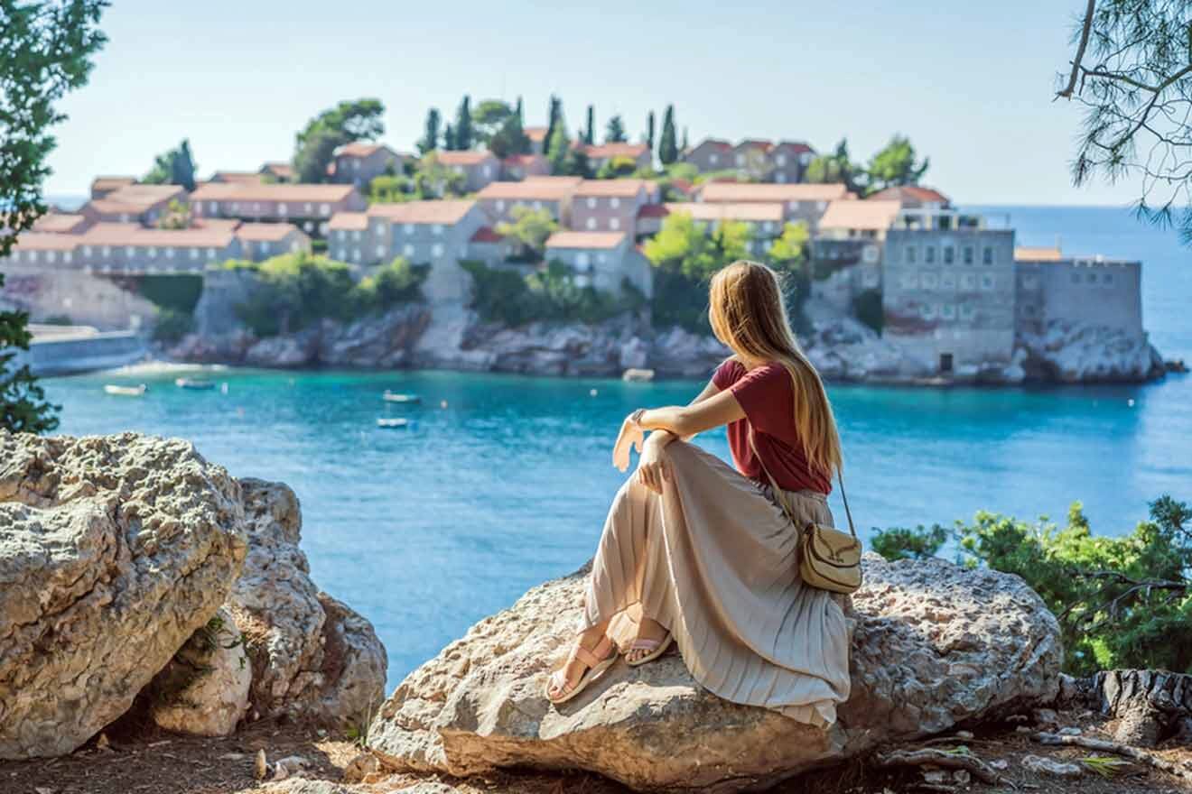 A woman sits on a rock overlooking a coastal town with red-roofed buildings surrounded by blue water and trees.