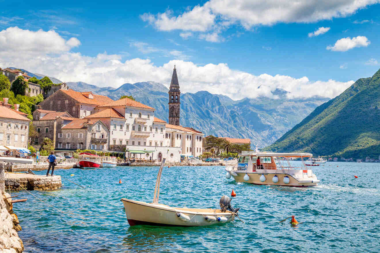 Coastal town with mountains, featuring a tall clock tower. Boats are docked in the water under a partly cloudy sky.