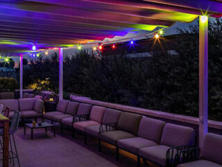 Outdoor terrace with a row of cushioned seating and a table, illuminated by colorful string lights under a pergola at dusk.