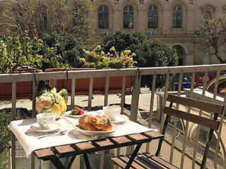 A small outdoor cafe table with two cups, a glass vase, pastries, and a bowl of fruit. There's a building and greenery in the background.