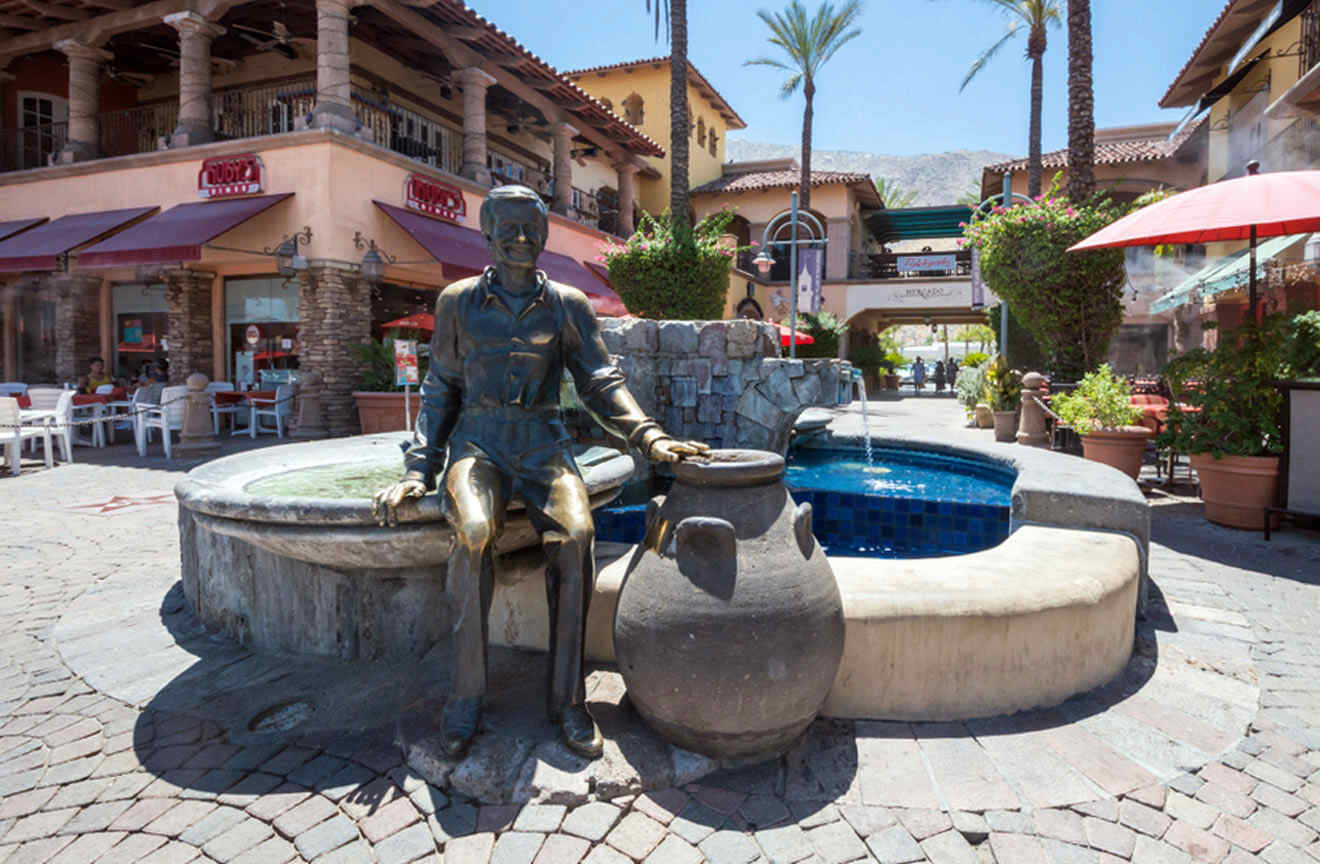 A statue of a man sitting on a fountain surrounded by shops and restaurants