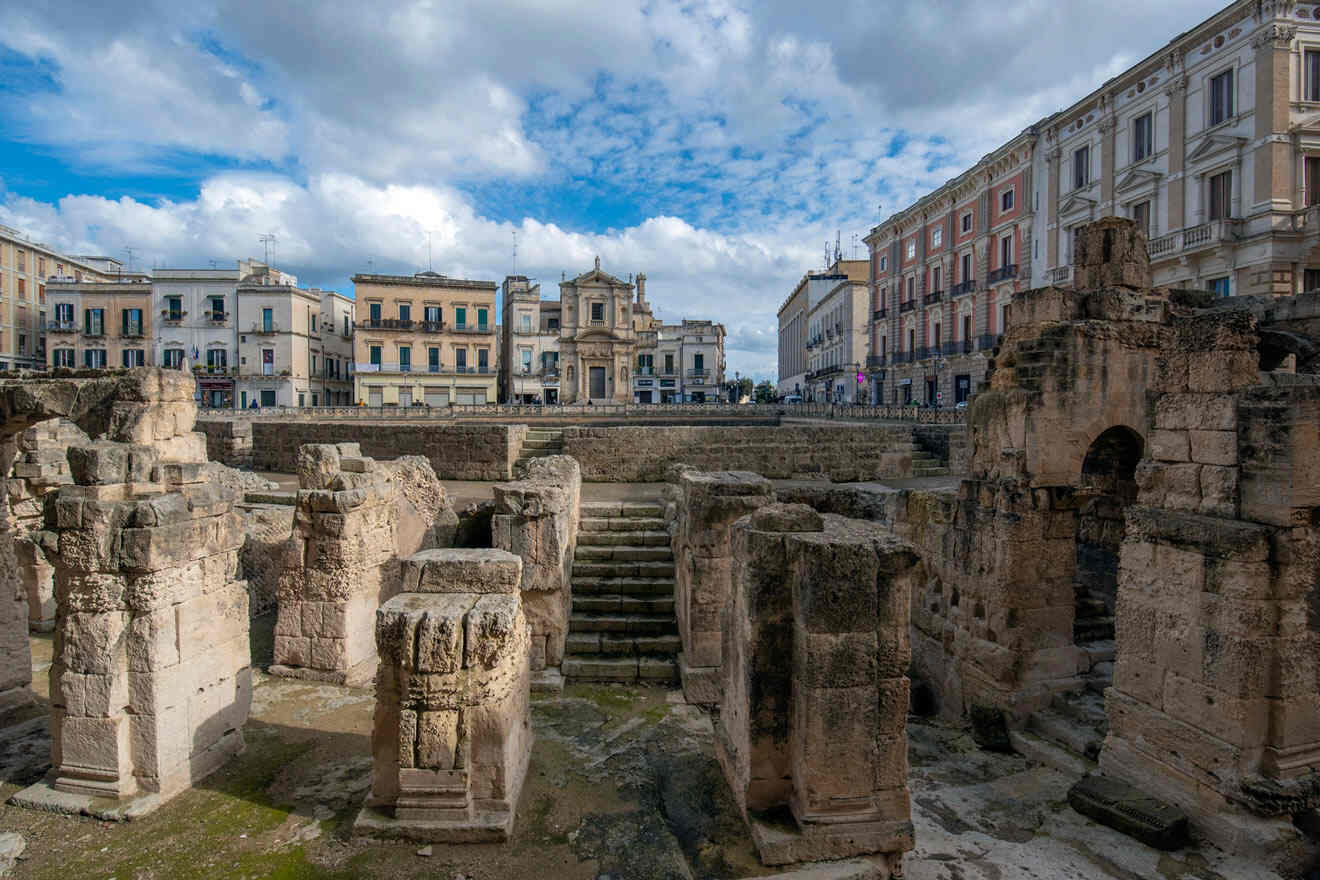 Ancient stone ruins in an urban square with surrounding historic buildings under a partly cloudy sky.