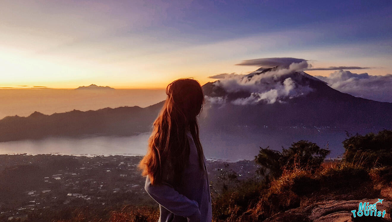 A woman is standing on top of a mountain looking at the sunset.