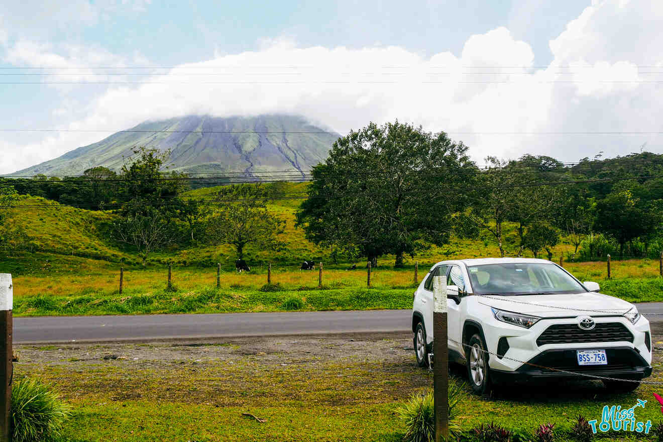 A white SUV is parked by the roadside near a grassy field, with a mountain in the background partially covered by clouds. The car has "665-726" on its license plate and a "Miss Tourists" sticker.