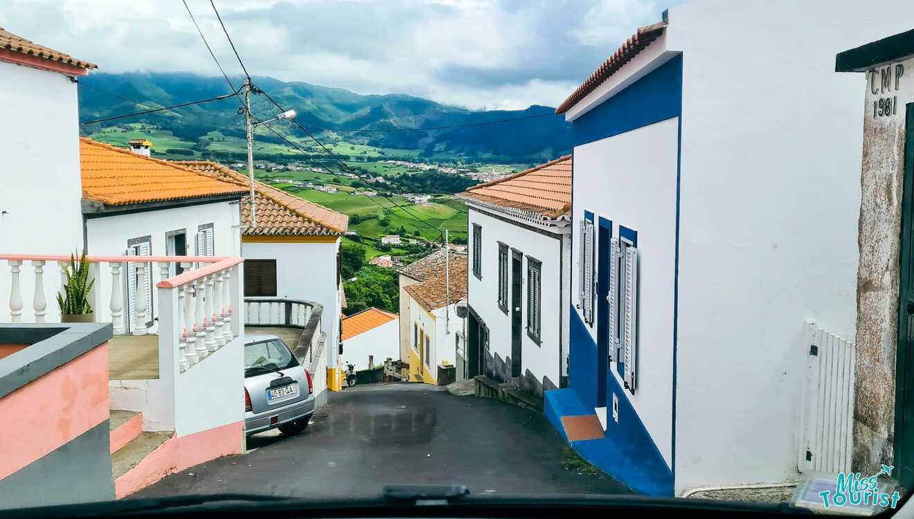 A car driving down a narrow street with houses and mountains in the background.
