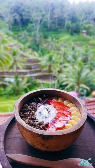 A bowl of fruit and yogurt on a table in front of a rice field.