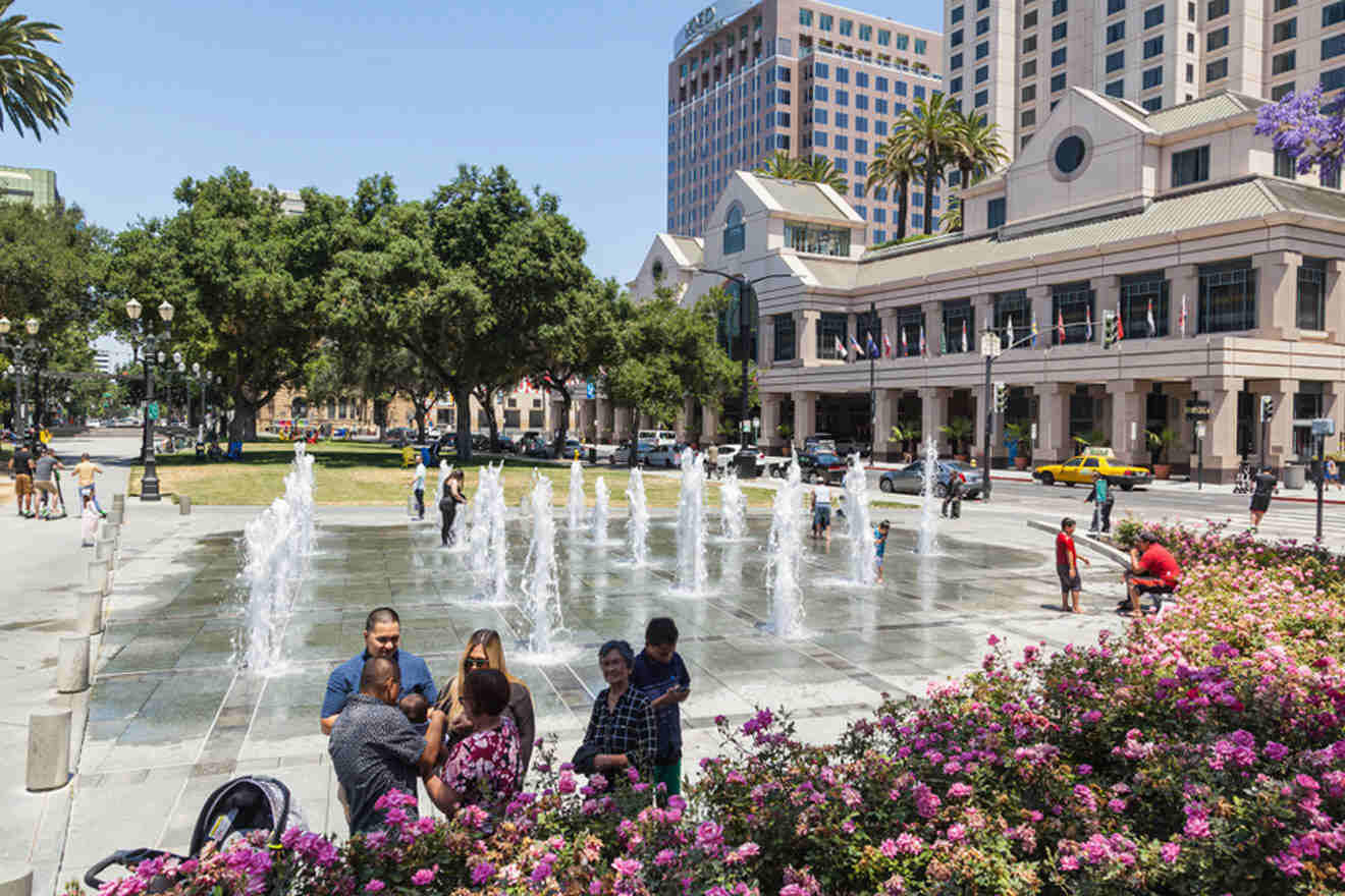 people and kids in the park with fountain and buildings in the background