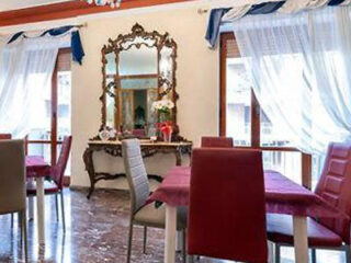 Dining area with several tables and chairs, an ornate mirror on the wall, and large windows with sheer curtains allowing natural light inside.