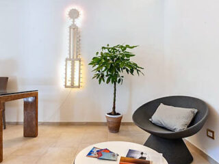 Modern interior with a potted plant, a black chair with a cushion, a small round table, and a sculpture-like wall light fixture.