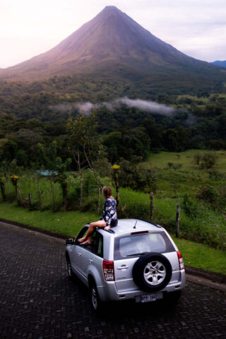 A person sitting on the roof of a car in front of a volcano.
