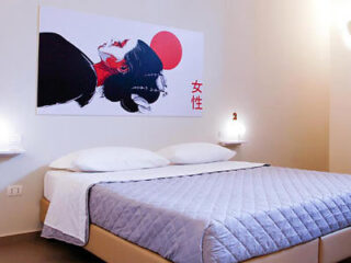 A minimalist bedroom with a double bed, gray quilted bedding, and artwork above featuring an abstract face and a red circle.