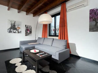 Modern living room with gray sectional sofa, black coffee table, round white lamp, orange curtains, wall art, and wood-paneled ceiling.