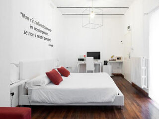 Minimalist bedroom with a white bed, red cushions, desk, and modern lighting. Italian quote on the wall.