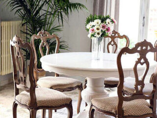 A round white table with four wooden chairs. A vase with flowers sits on the table. A potted plant is in the background.