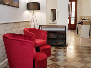 A room with two red chairs and a small table on a tiled floor. A floor lamp stands nearby, and a doorway is visible in the background.