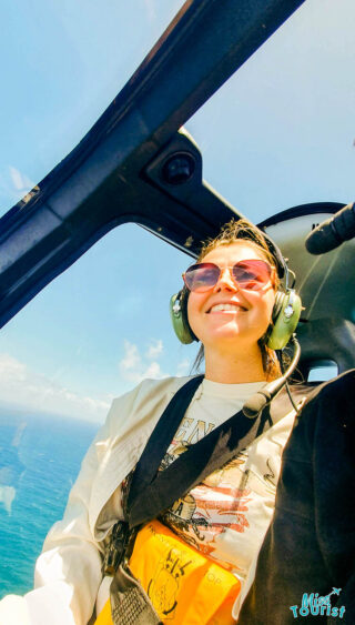 author of the post smiling inside a helicopter, wearing sunglasses, a headset, and a safety harness. Ocean and sky visible through windows.