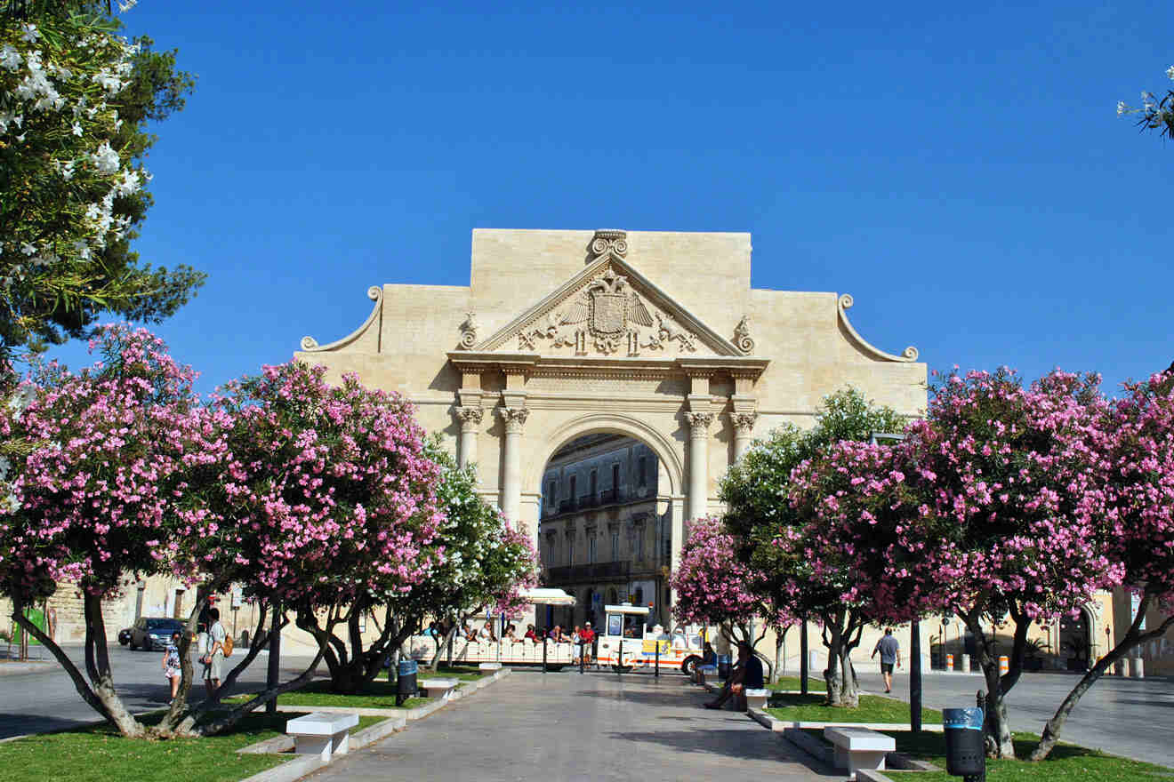 A large stone archway with decorative carvings stands as a focal point, surrounded by blooming pink trees and benches. People walk nearby under a clear blue sky.