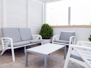 Outdoor patio with white plastic furniture, including a sofa, two chairs, a low table, and potted plants, on a tiled floor.