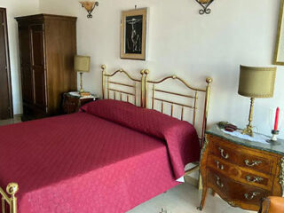 A bedroom with a brass bed frame and red bedspread, wooden furniture, two lamps, and a wall painting above the bed.