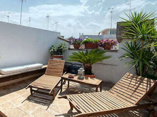 A rooftop patio with two wooden lounge chairs, a small table, potted plants, and a cushioned bench under a cloudy sky.