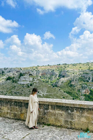 A woman in a white robe standing near the small stone wall.