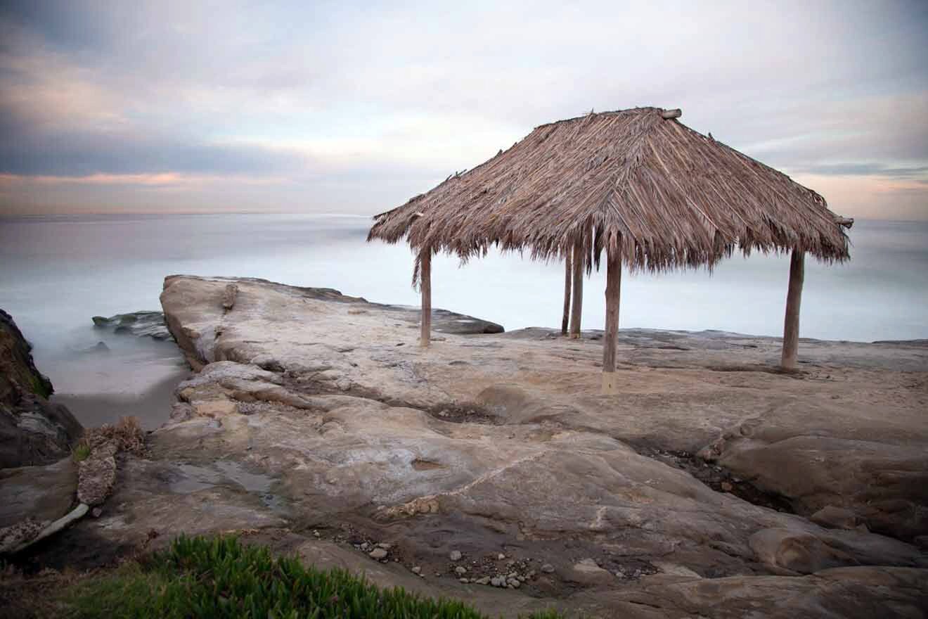 A thatched hut sits on top of a rock next to the ocean.