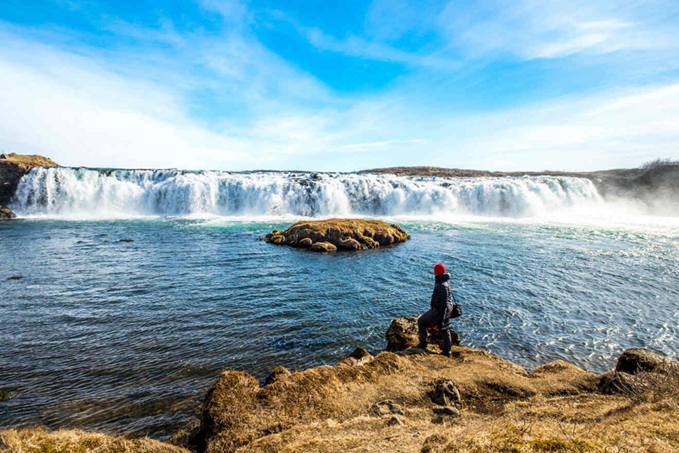 Person in a red hat sitting on a rocky outcrop, gazing at a majestic waterfall in a serene landscape, with mist rising from the turbulent water below