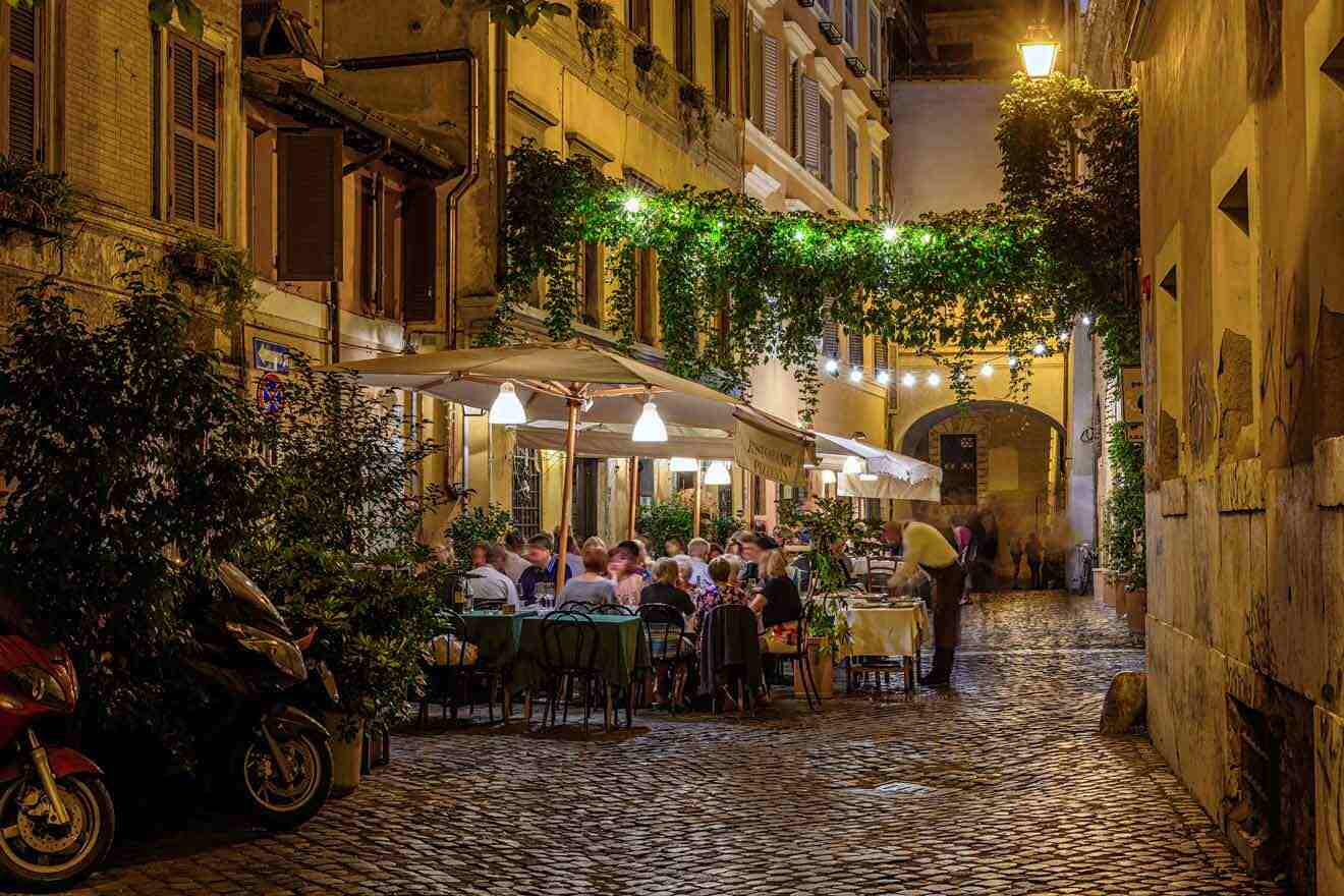 A narrow cobblestone street at night with people sitting at tables.