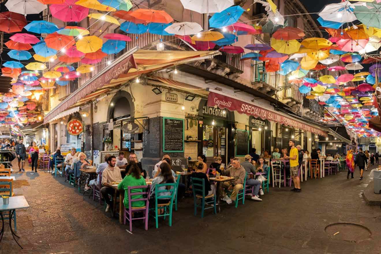 Colorful umbrellas hanging from the ceiling of a street with people sitting at an outdoor restaurant