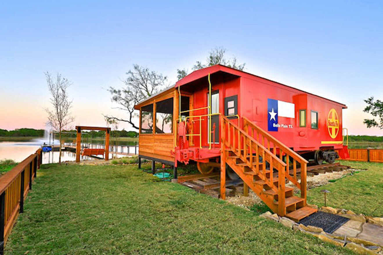 A red train cabin sits on a lawn next to a lake.