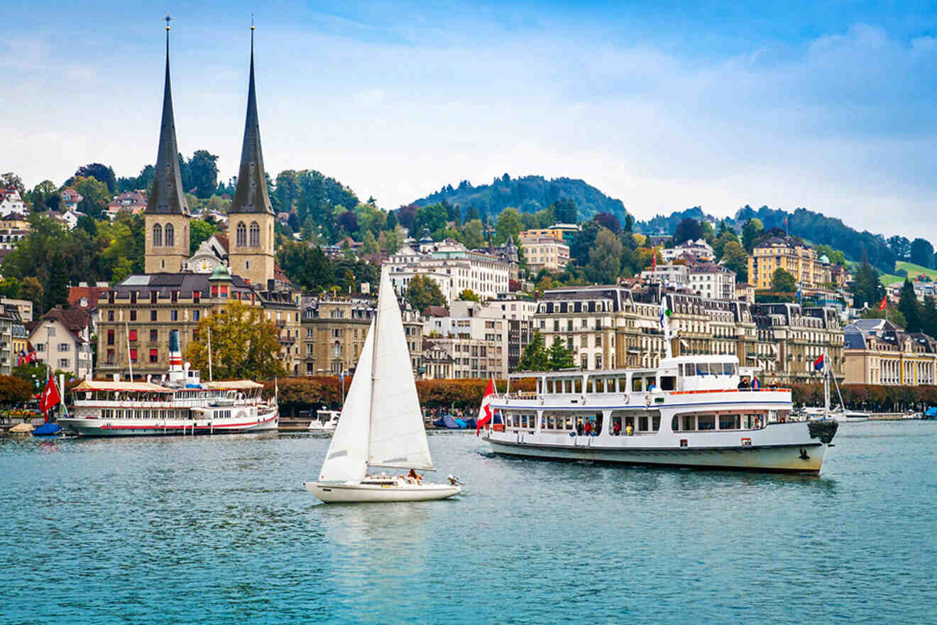 Several boats, including a sailboat and passenger ferries, sail on a lake with a scenic town and twin-spired church in the background. Hills and buildings frame the waterscape under a partly cloudy sky.