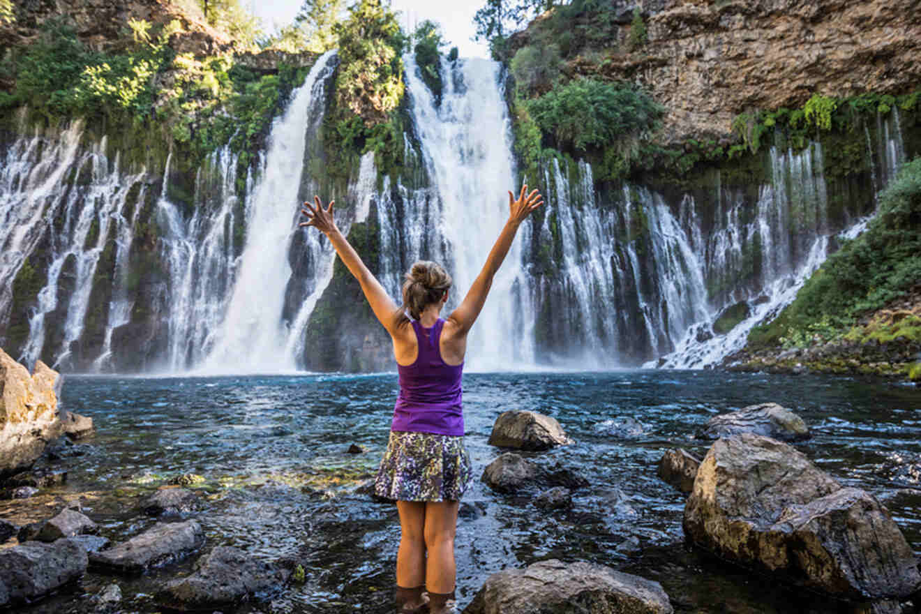 A woman is standing in front of a waterfall with her arms raised.
