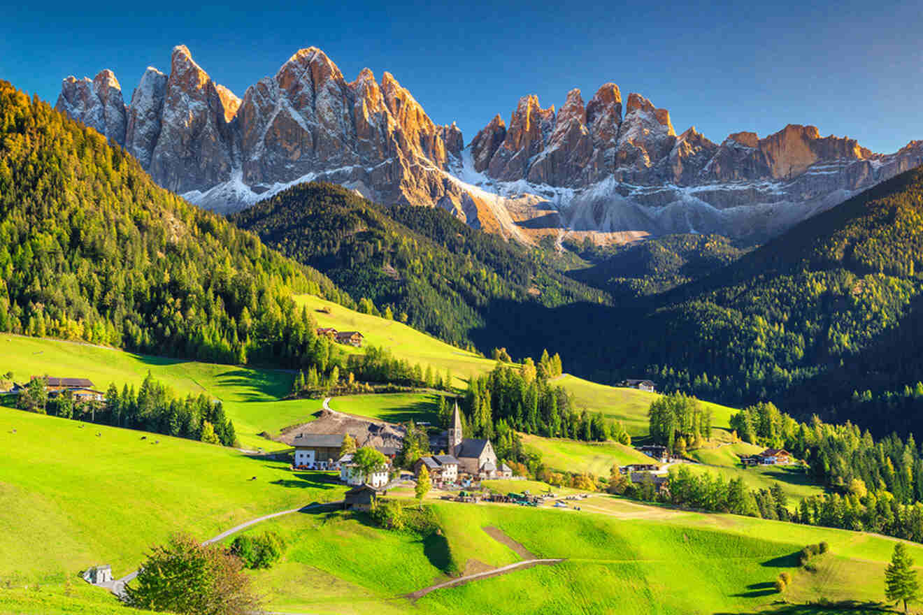 A picturesque village is nestled in a verdant valley with sunlit green hills and tall jagged mountains in the background under a clear blue sky.