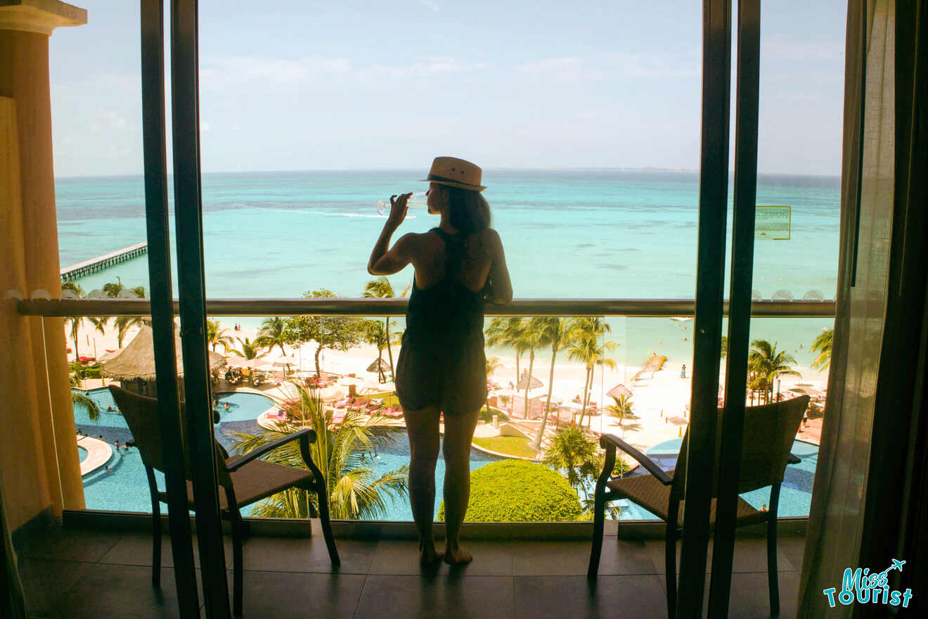 A woman with a glass of champagne in a hat looking out of a window at the beach.