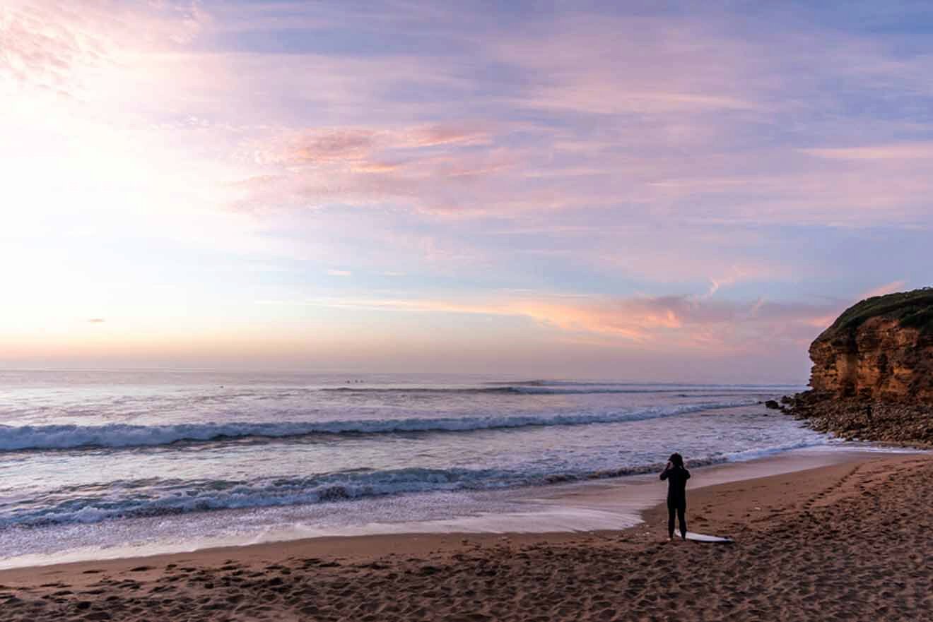 A surfer is standing on the beach at sunset.