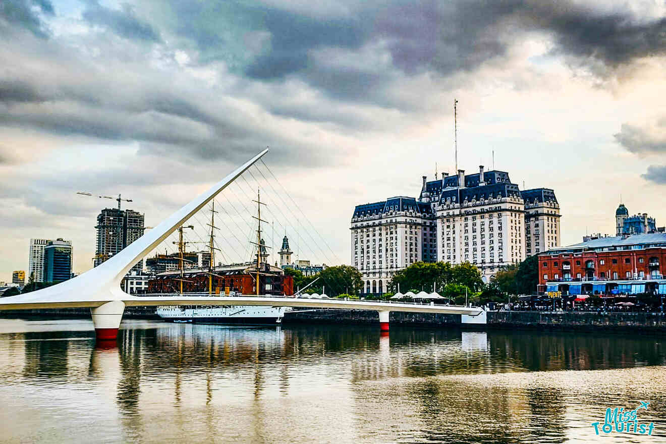 The iconic Puente de la Mujer, a modern pedestrian bridge in Buenos Aires, with a moored ship and historic buildings, against a dramatic cloudy sky at twilight