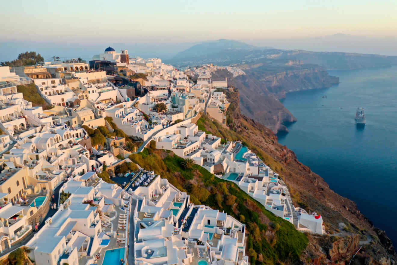 Aerial view of Santorini at dusk, showcasing the clustered white buildings and pools along the caldera edge with a cruise ship in the distant sea