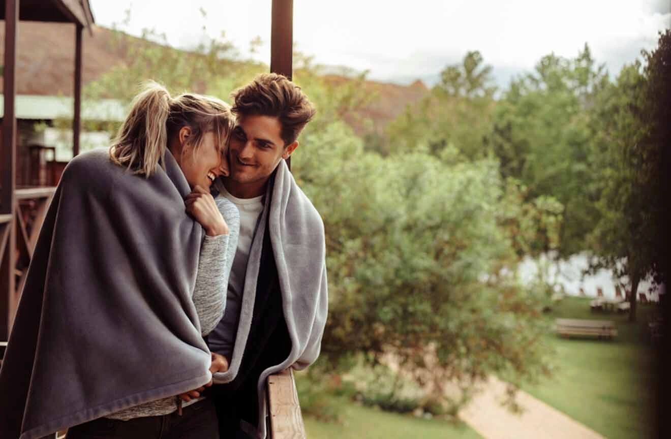 A couple wrapped in blankets on a deck overlooking a lake.