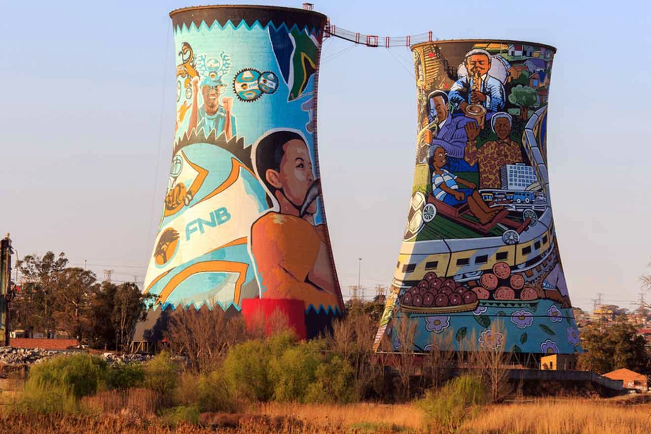 Two large cooling towers adorned with vibrant murals depicting various cultural and historical scenes. The towers stand in a grassy field with some industrial structures visible in the background.