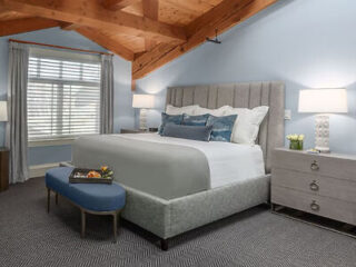 A bedroom with a gray upholstered bed, blue bench, two nightstands with lamps, and a window with white blinds under a wooden beamed ceiling.
