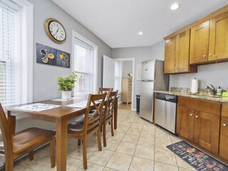 A kitchen with wooden cabinets, stainless steel appliances, a wooden dining table with four chairs, tiled floor, and wall clock. A plant and artwork are on the table and wall.