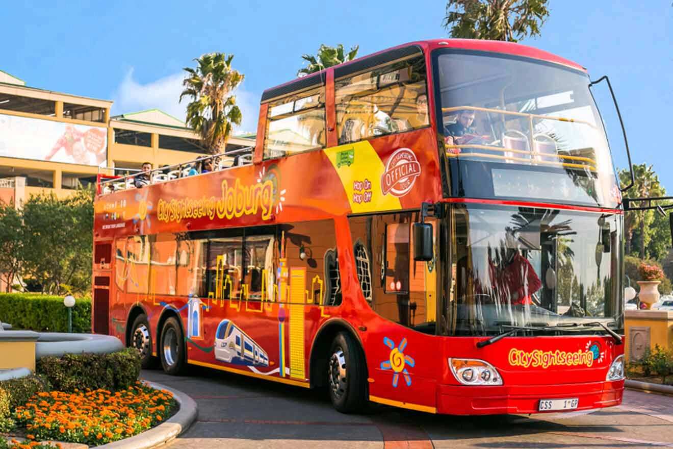 A red double-decker tour bus with sightseeing graphics and branding, parked near a landscaped area with palm trees and flowers.