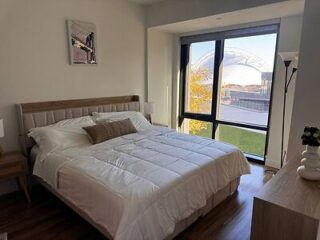 A neatly made bed with white bedding in a modern bedroom, featuring a large window with a view of an outdoor structure and greenery.