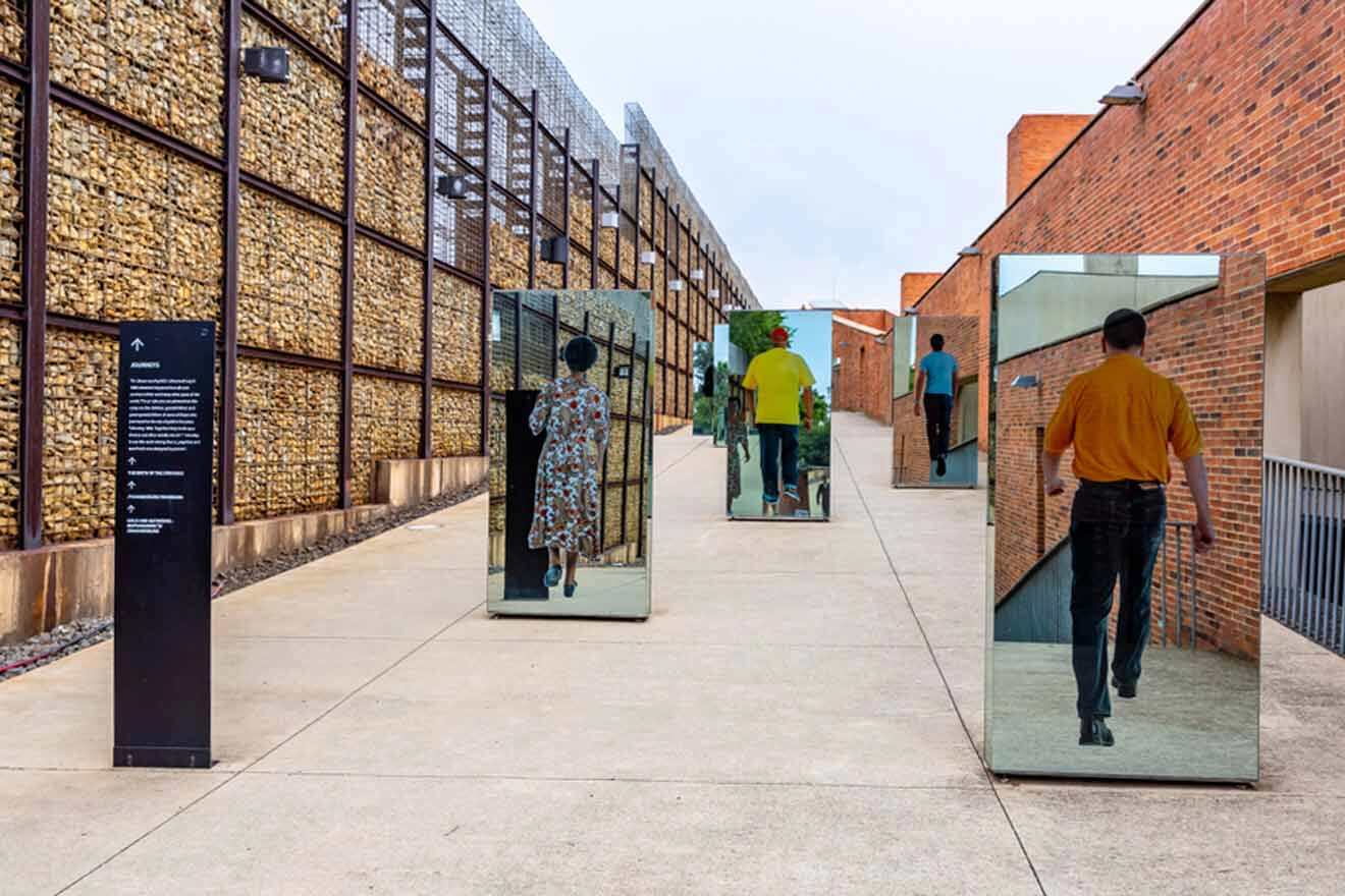 Outdoor art installation with multiple mirrors reflecting people walking. The mirrors are placed along a pathway between two brick and wire-mesh walls.